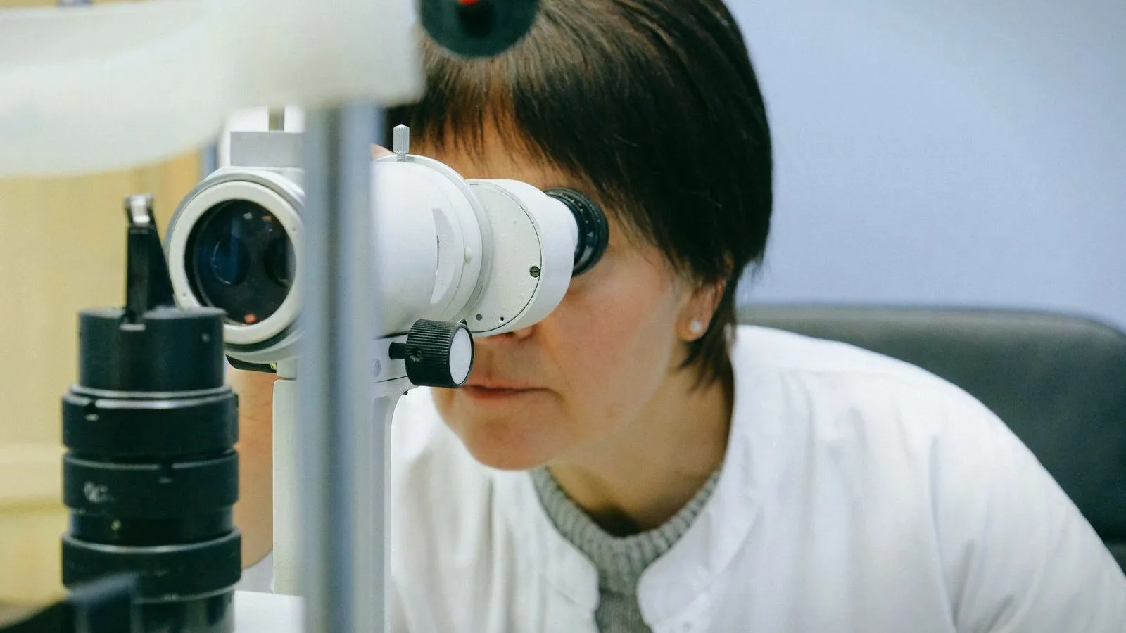 A clinician in a white coat looking through an eye examination device in a clinic