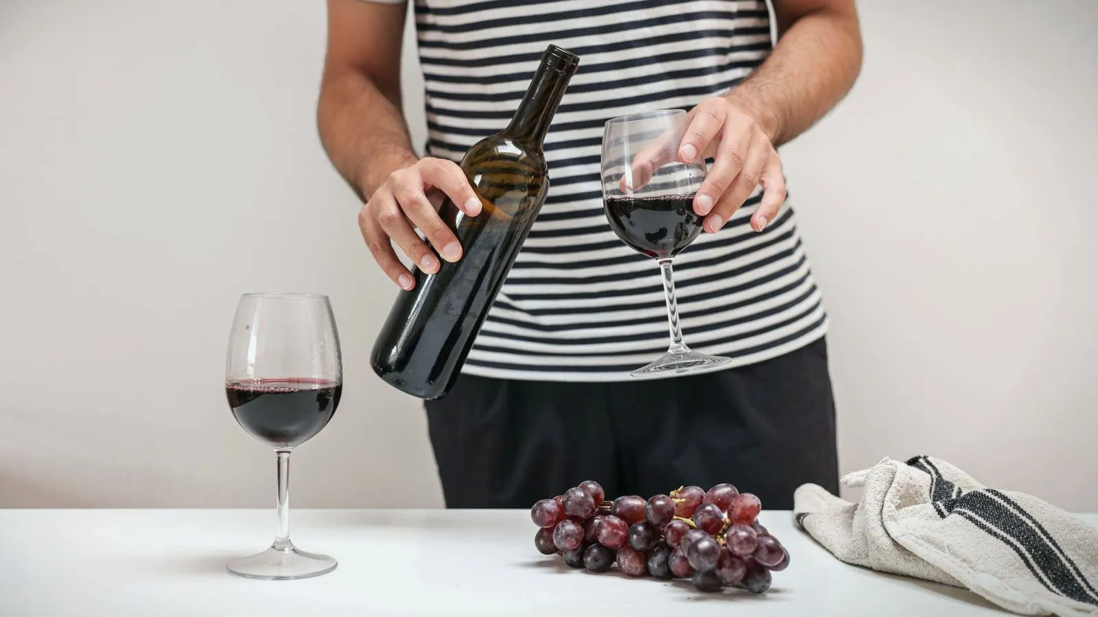A person pouring red wine into a glass beside a bunch of grapes
