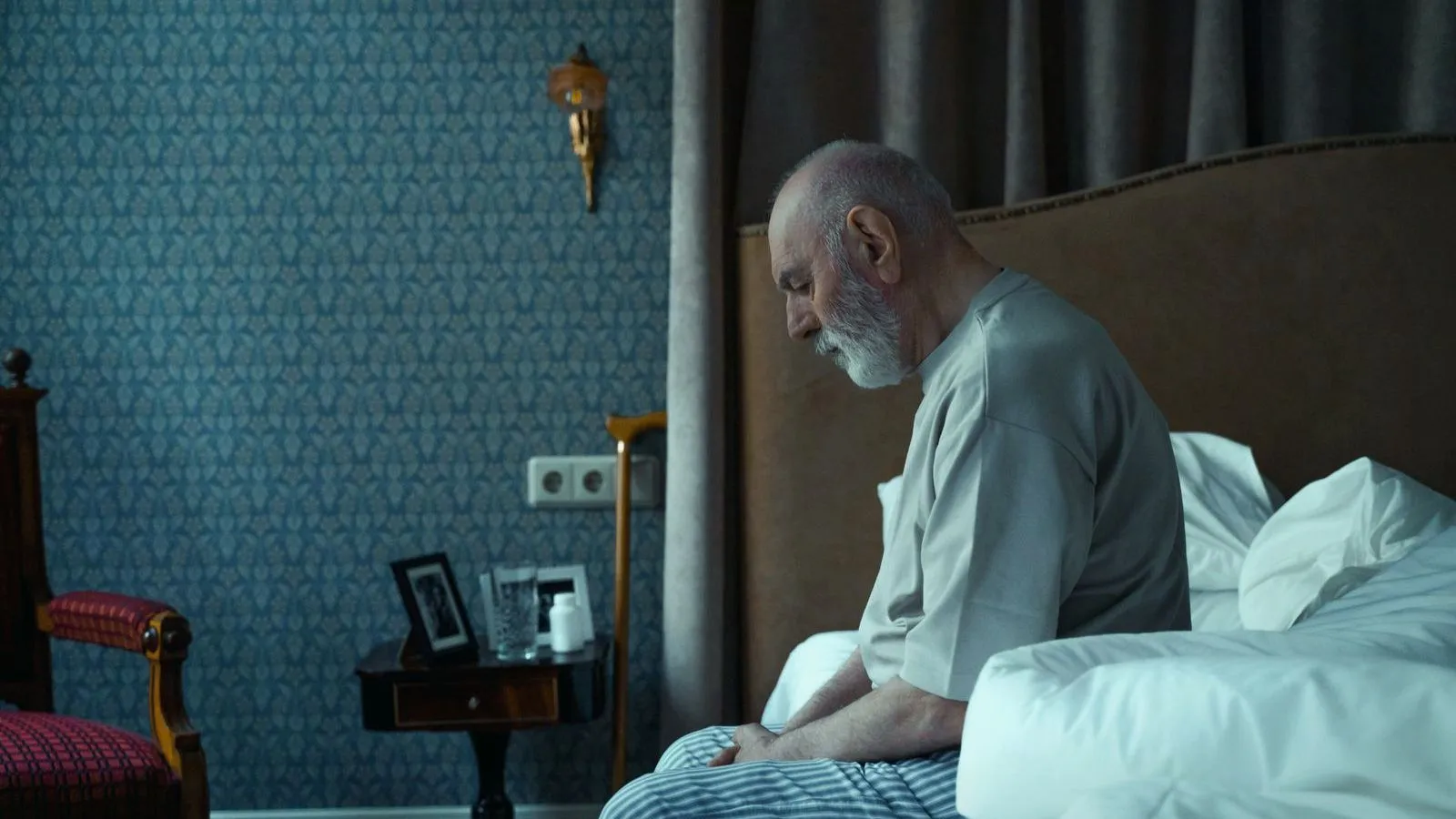 An older man sitting on the edge of a bed with his head lowered