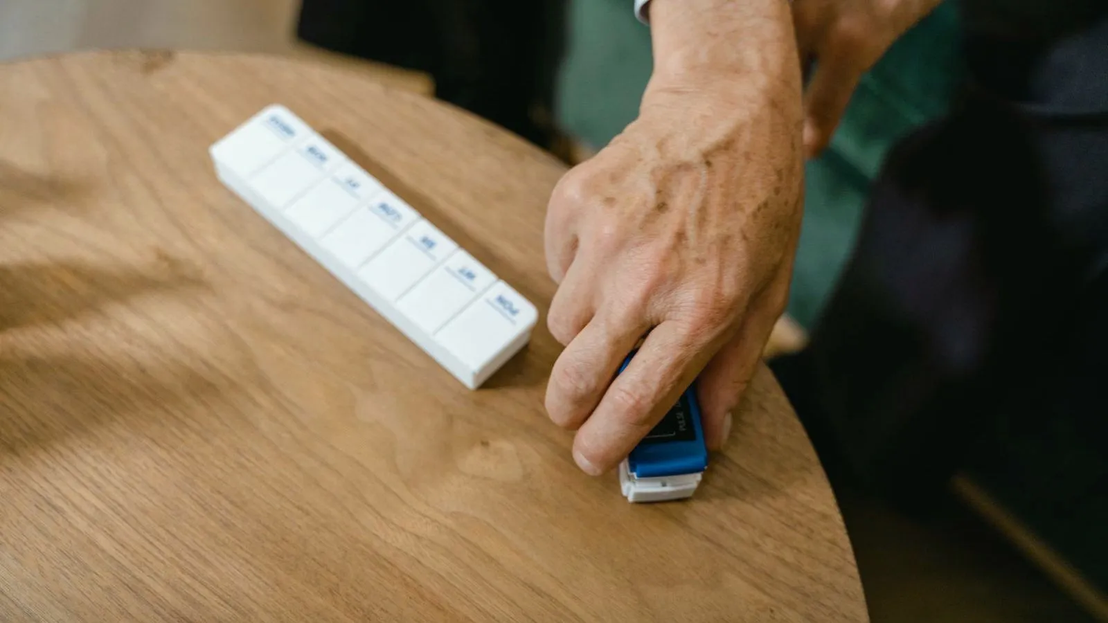 An older person's hand reaching for a weekly pill organizer on a wooden table