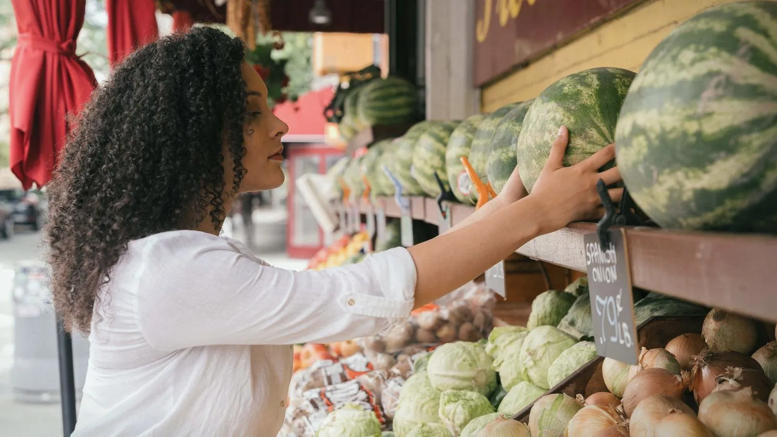 A shopper reaching for a watermelon at an outdoor produce stall with chalked prices