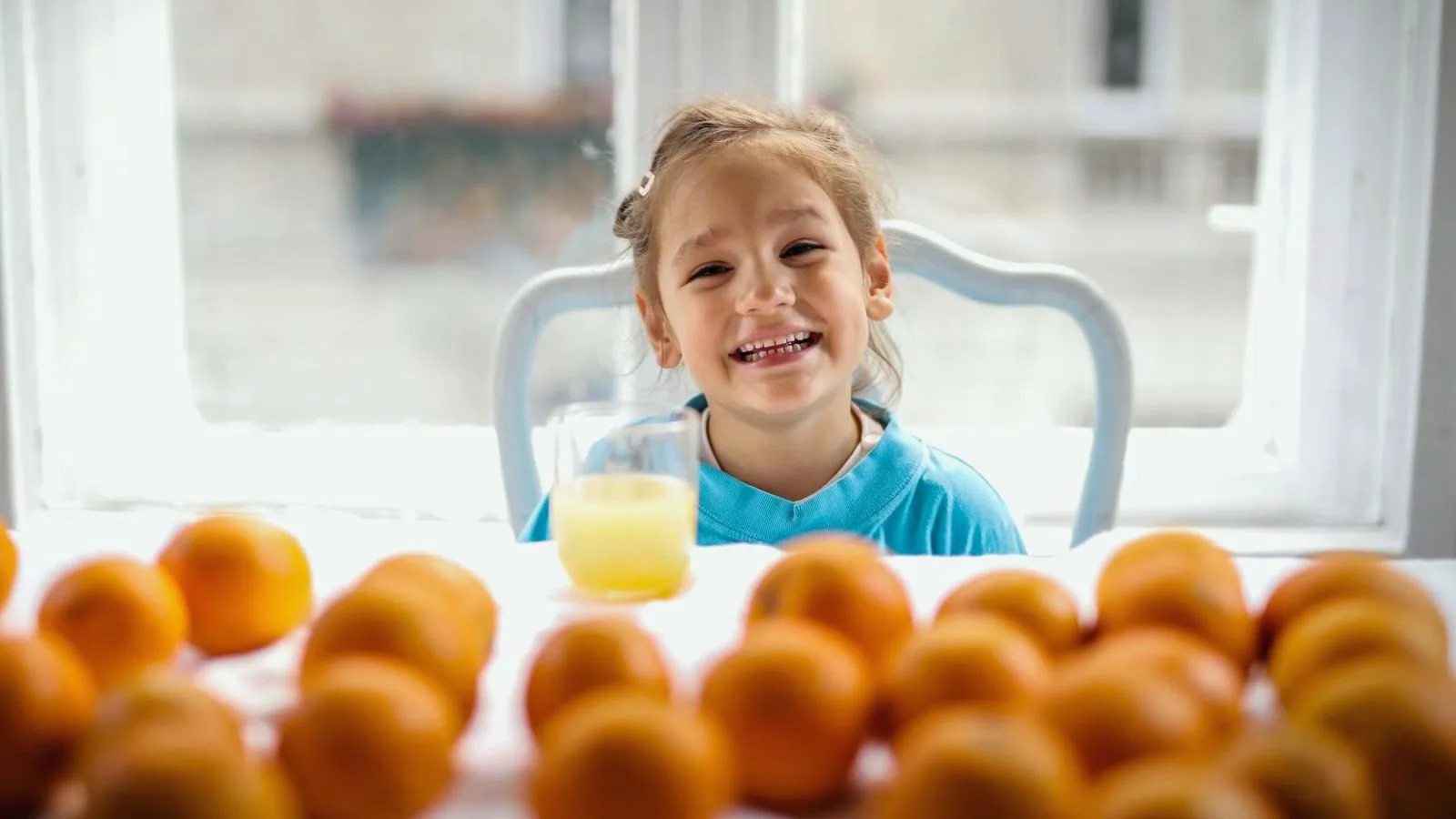 A young girl smiling at a table behind a glass of juice, with whole oranges in the foreground