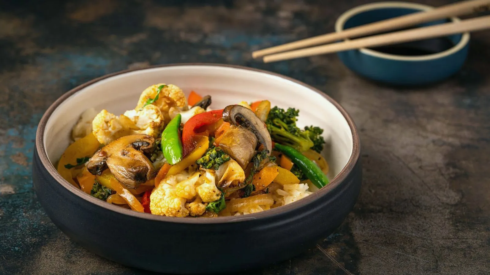 A bowl of stir-fried vegetables over rice with chopsticks resting alongside