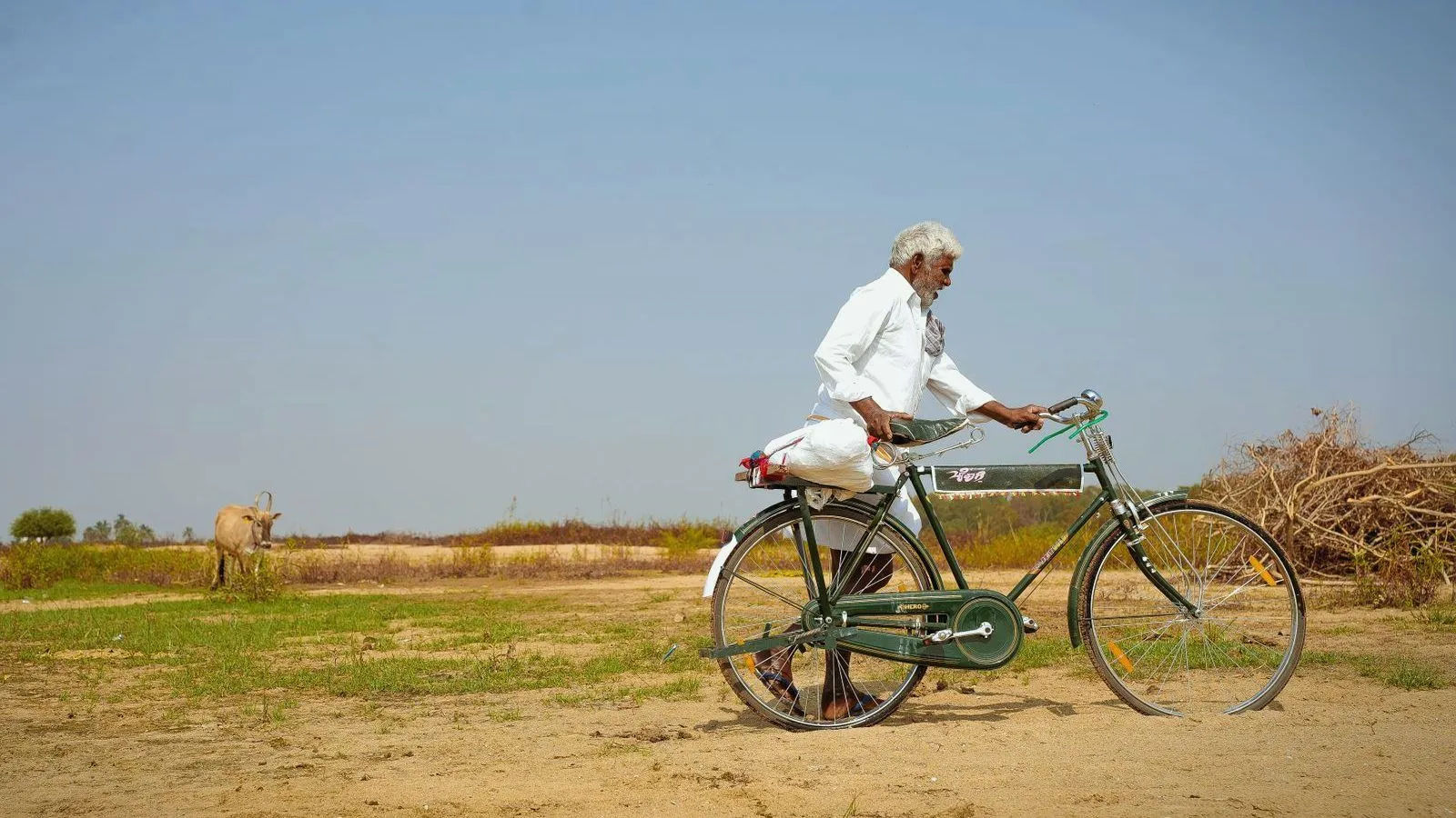 An older man in white clothes walking a bicycle along a sandy rural path in India