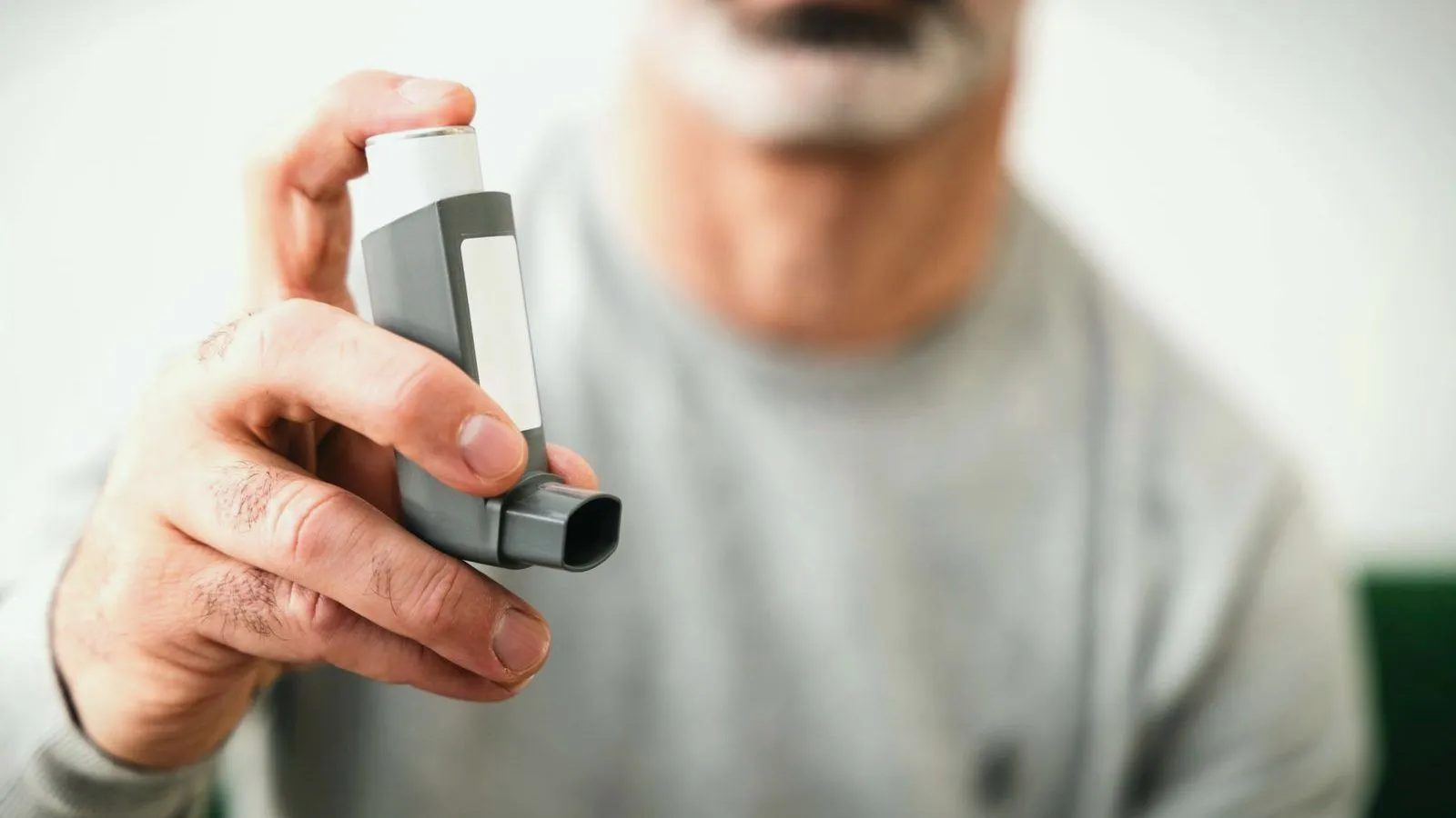 An older man with a grey beard holding an inhaler, his face out of focus behind it
