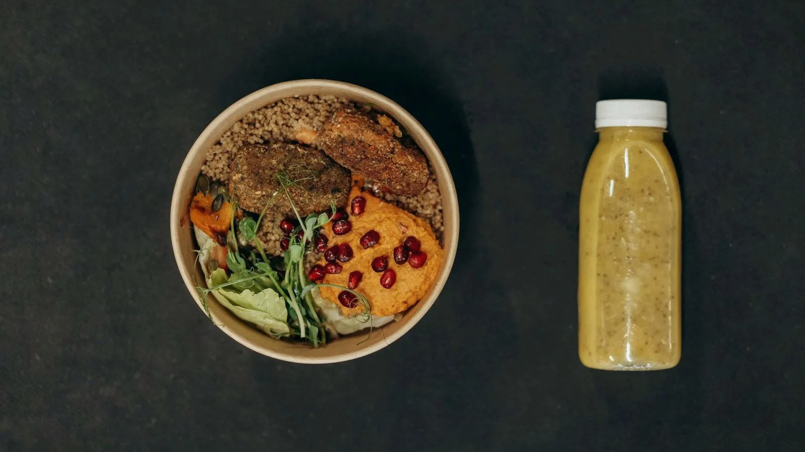 An overhead view of a bowl of whole food beside a bottled liquid drink on a dark surface