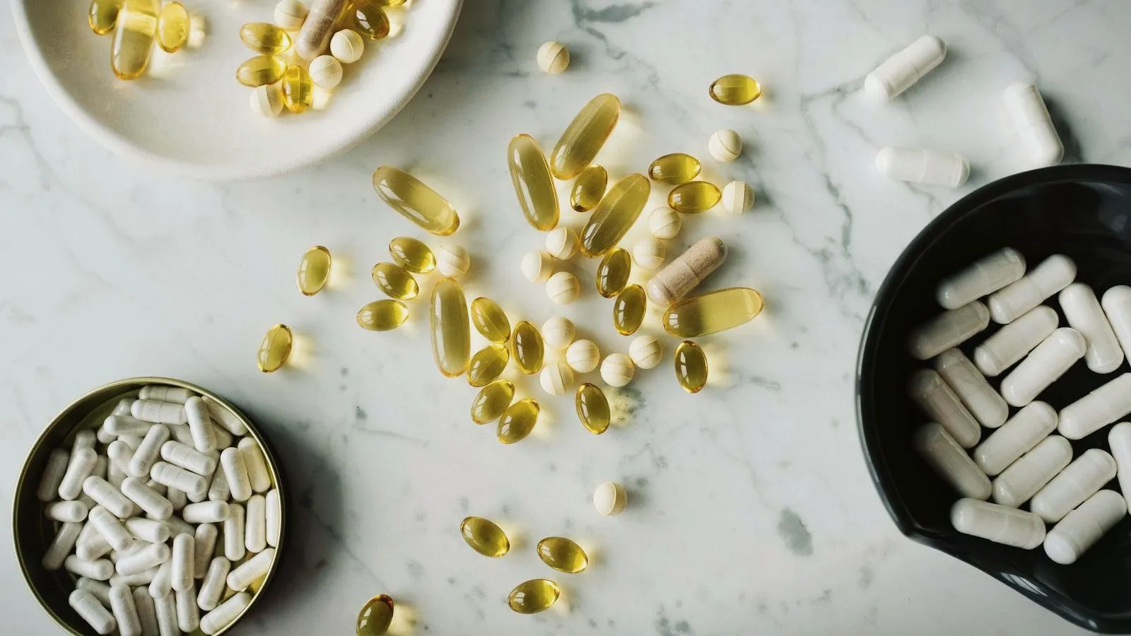 Softgel and capsule supplements scattered across a marble surface beside small bowls