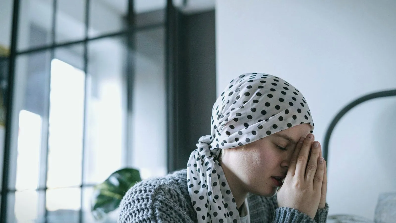 A woman in a spotted headscarf and grey cardigan sitting with her eyes closed and her hands pressed to her face