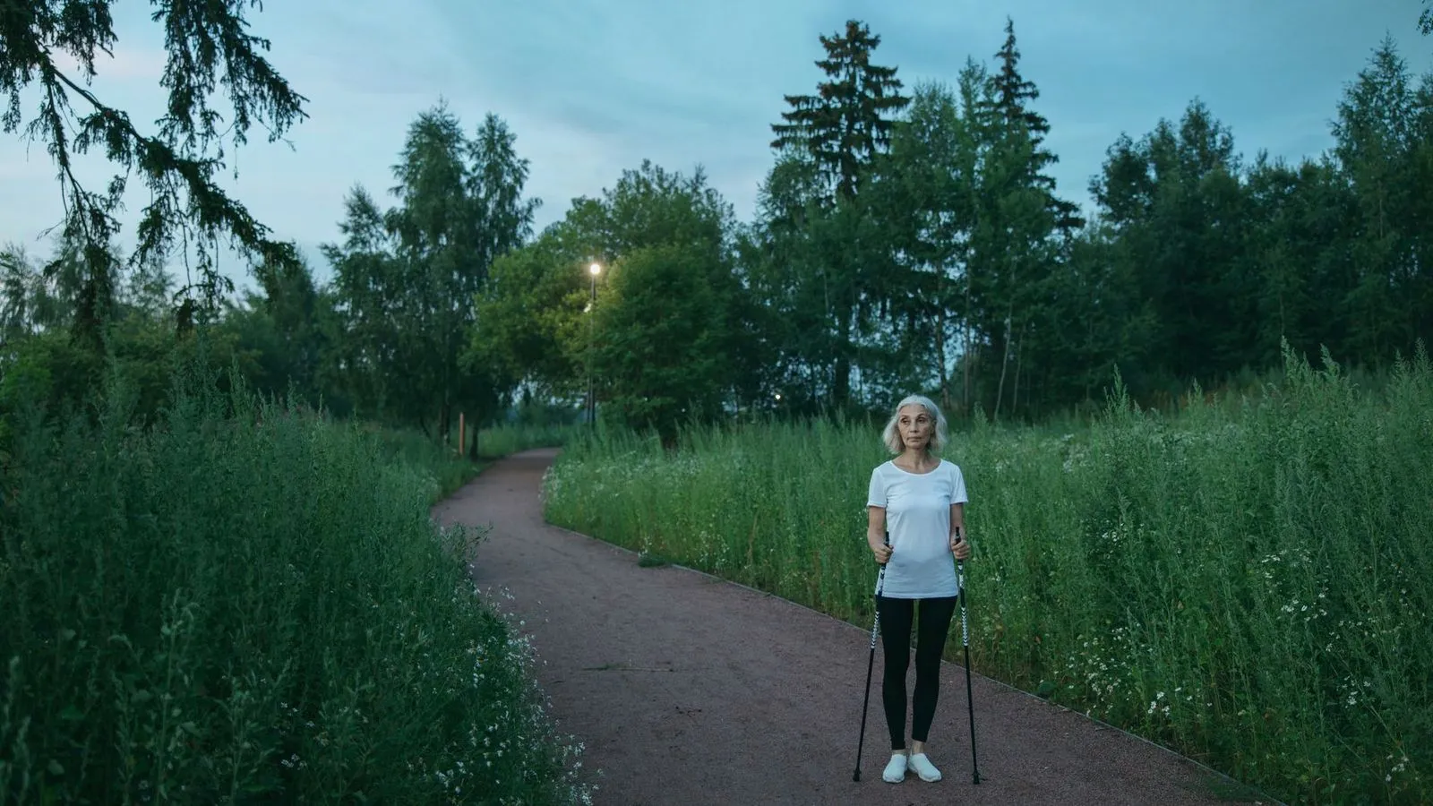 An older woman in athletic wear walks a park path with Nordic walking poles at dusk