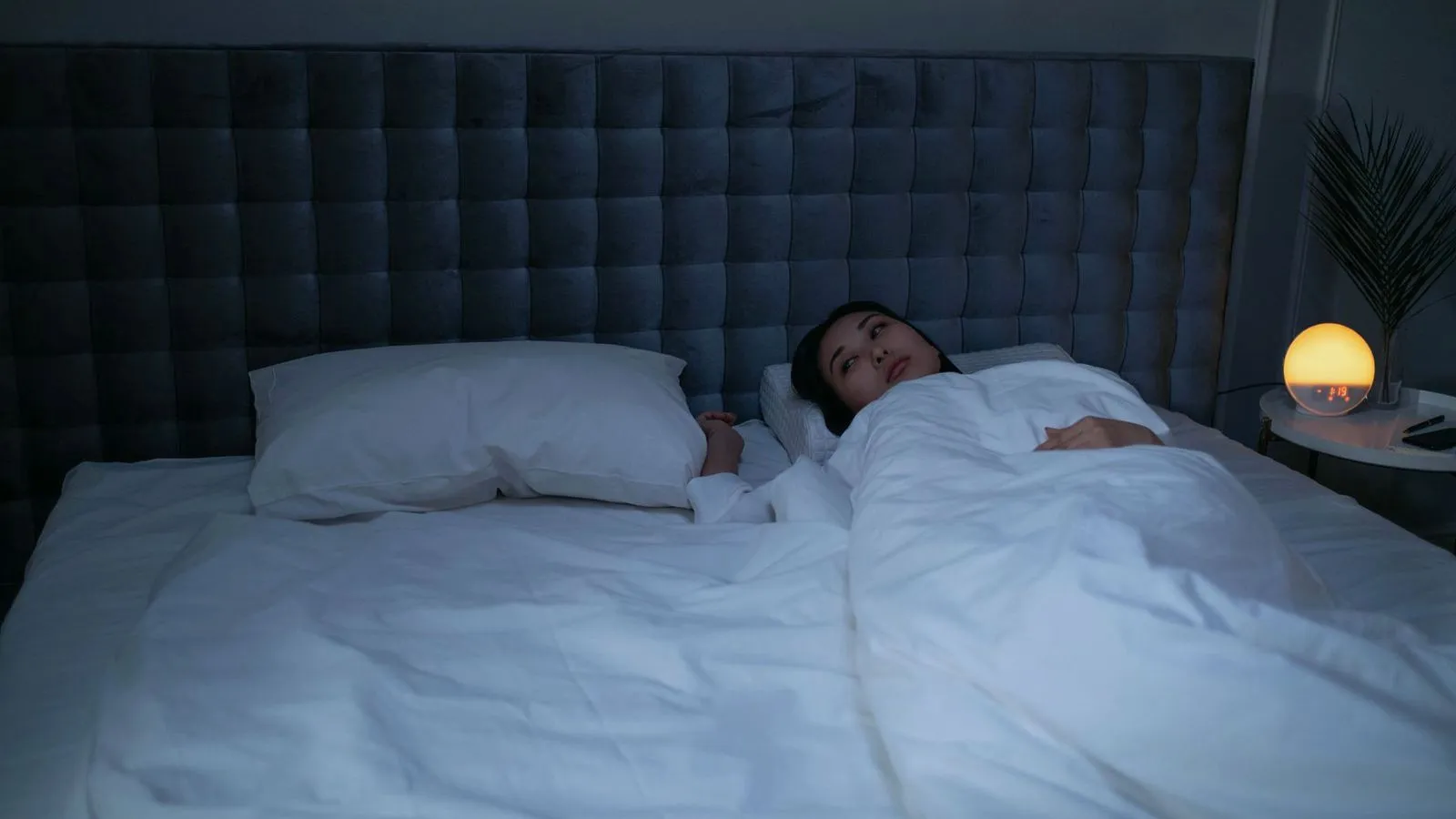 A woman lying awake in bed at night with a bedside lamp glowing