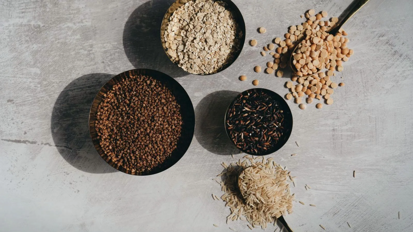 Four dark bowls holding oats, buckwheat, wild rice and split peas, with rice and lentils spilling from brass spoons on a pale stone surface