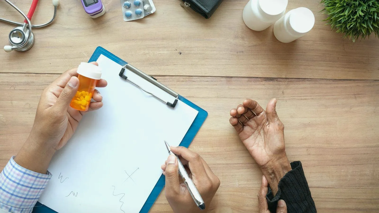 An overhead view of a clinician holding a pill bottle and pen over a clipboard, across a desk from an older patient's hands