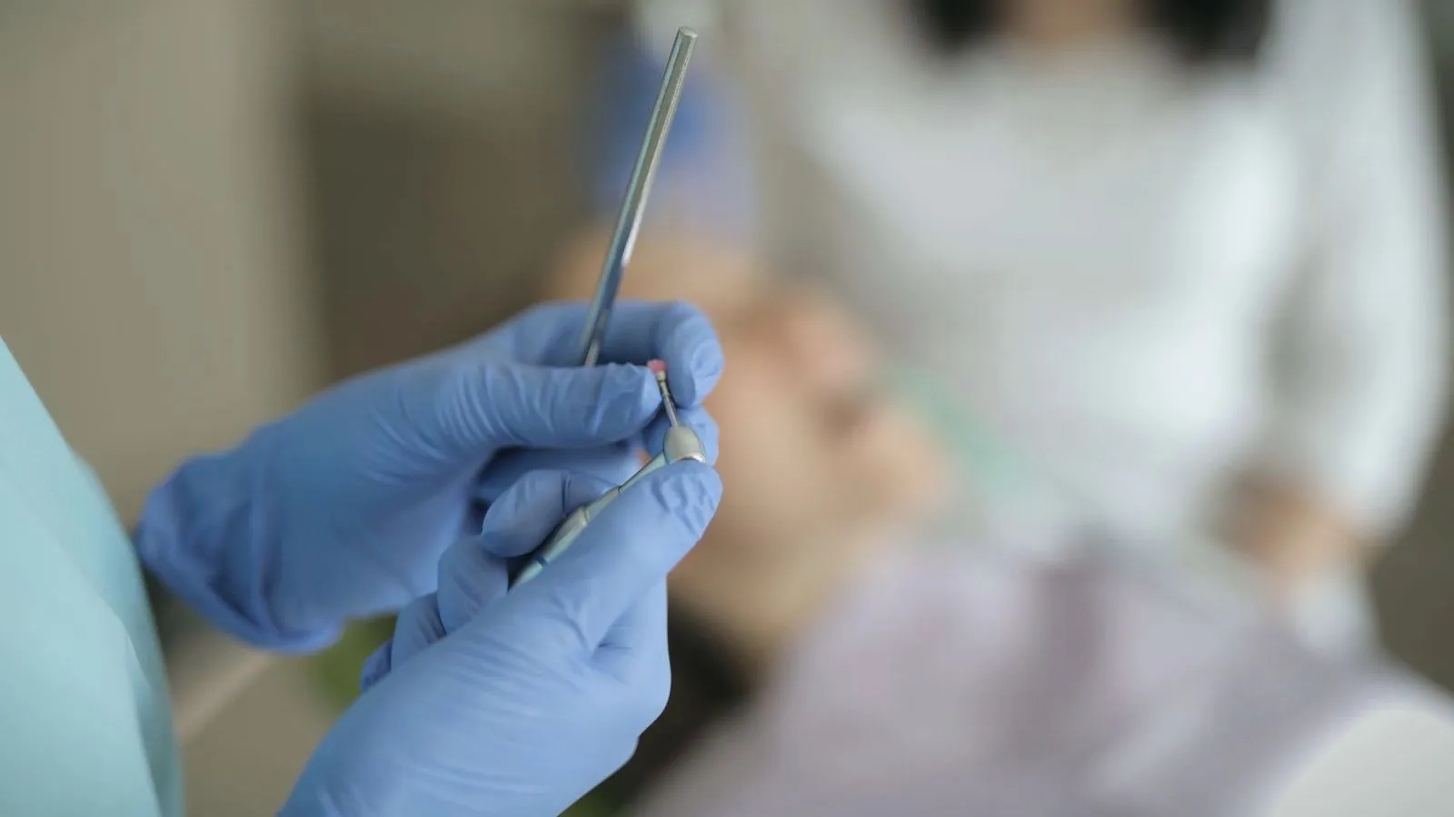 A gloved hand holding a dental instrument, with a reclining patient blurred behind