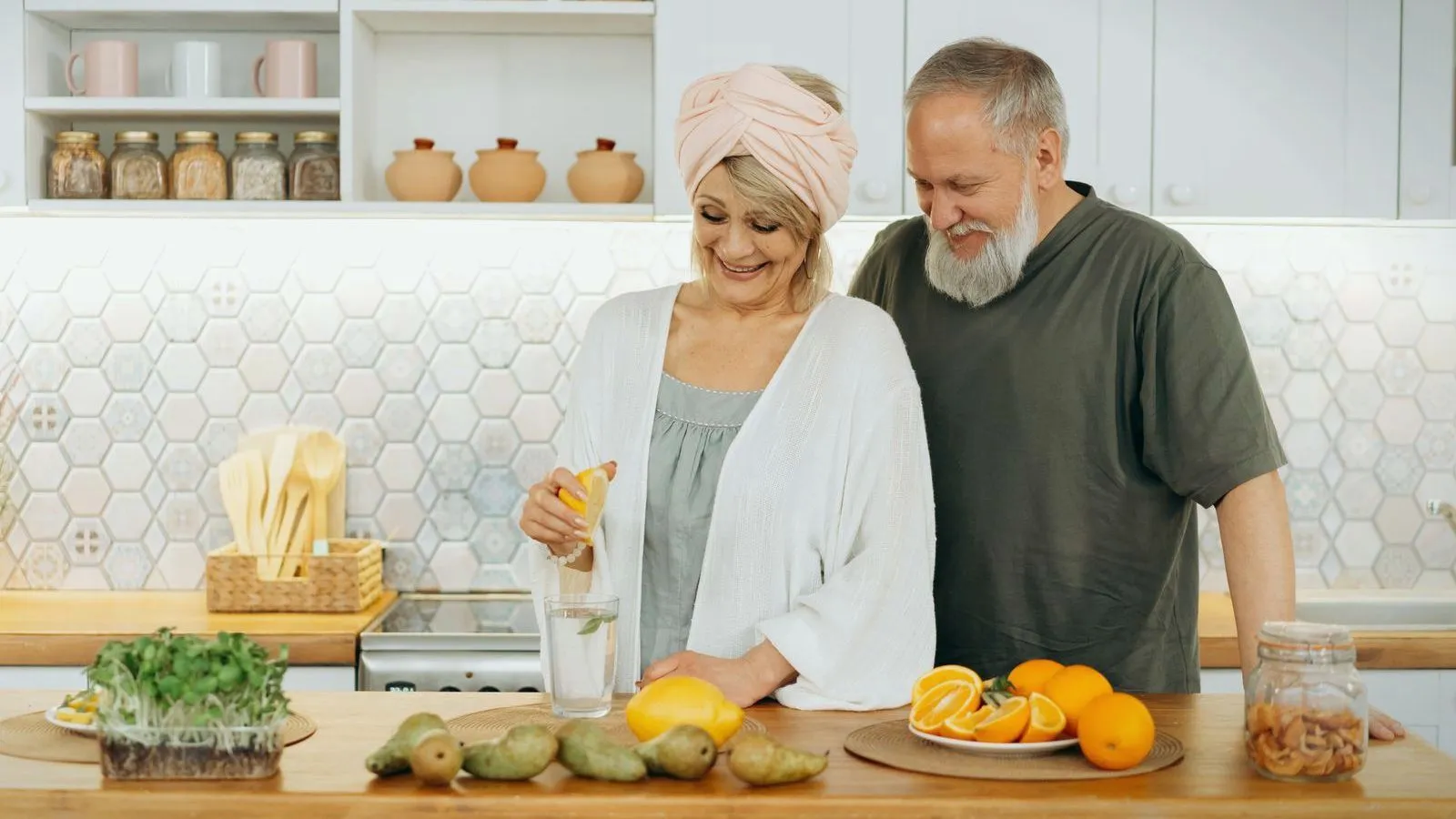 An older couple in a bright kitchen with pears, oranges and lemons on the counter