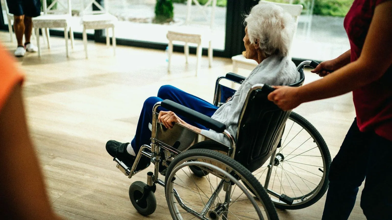 An elderly woman being pushed in a wheelchair along a bright care home corridor