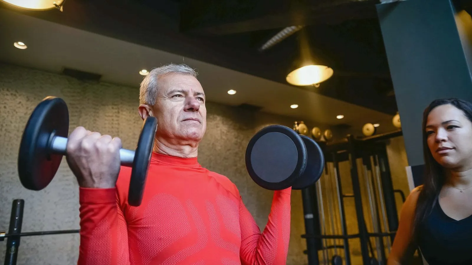 An older man lifting dumbbells in a gym with a trainer nearby