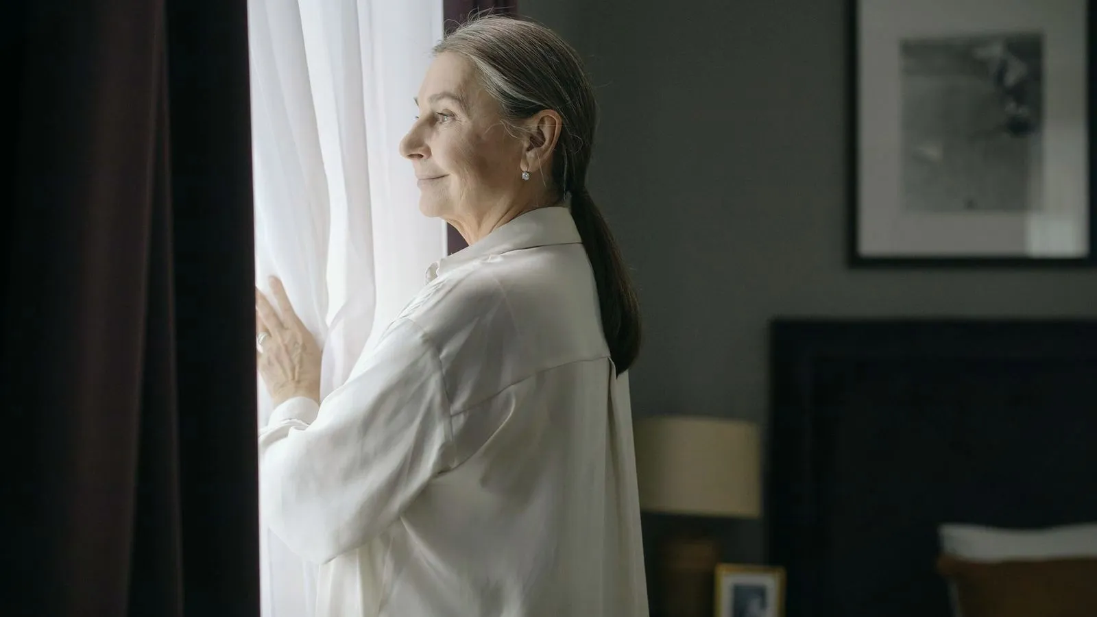 An older woman standing alone at a window, holding the sheer curtain and gazing out
