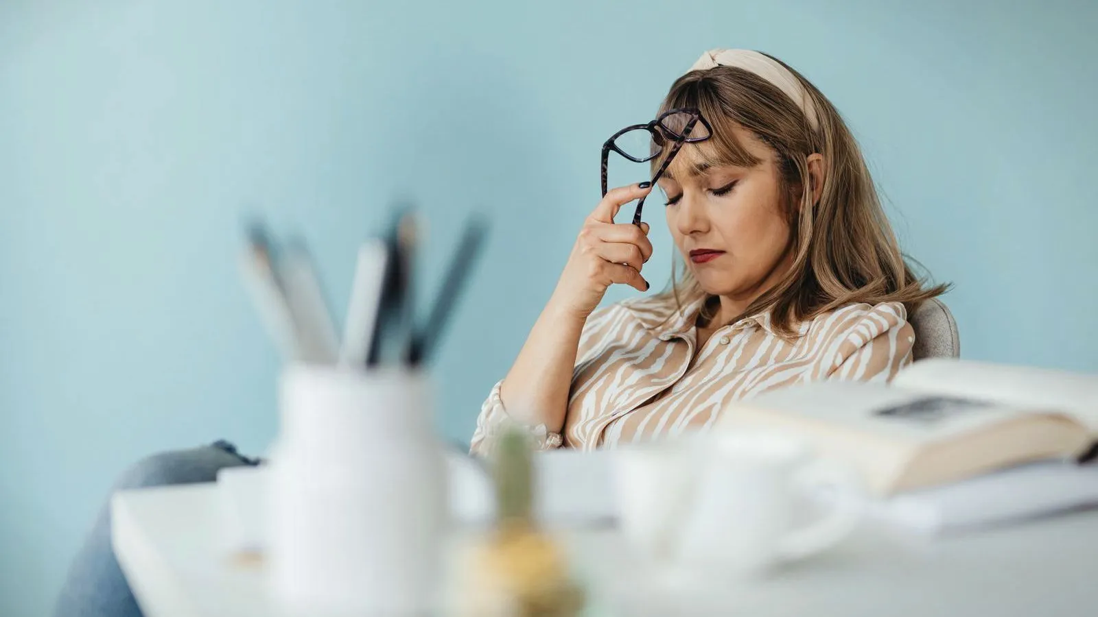 A woman at a desk lifting her glasses and closing her eyes over an open book