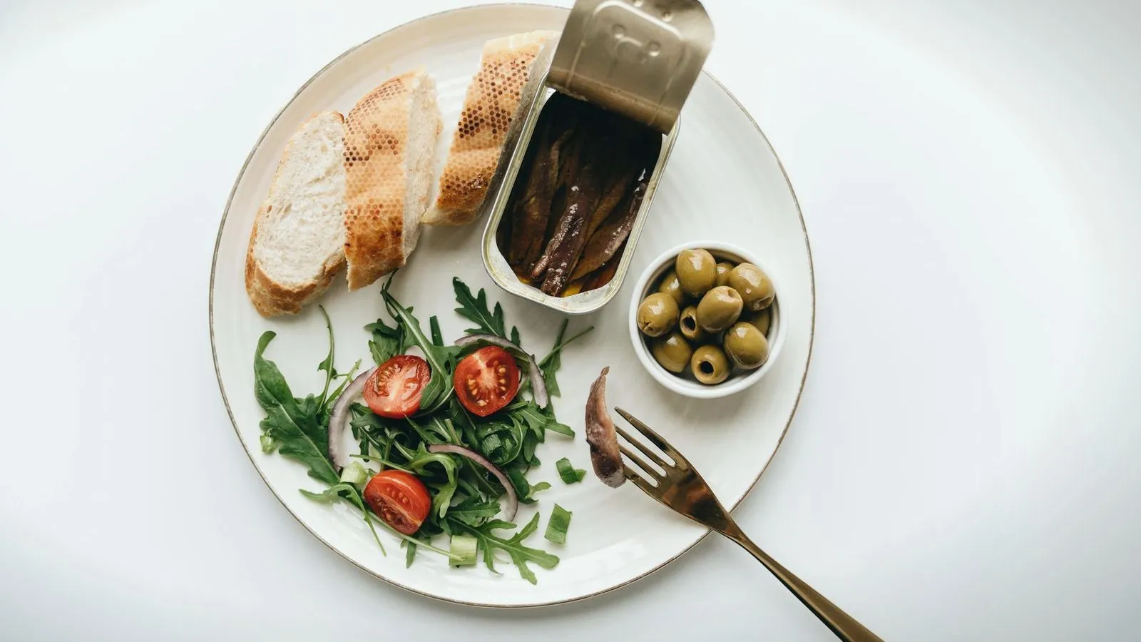 An overhead plate of bread, anchovies, olives and salad leaves