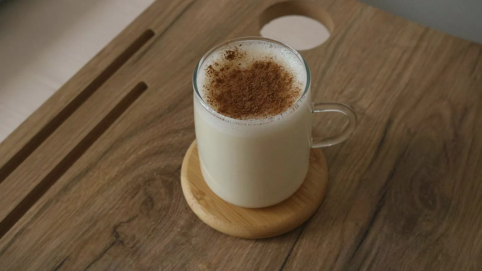 A glass mug of warm milk dusted with cinnamon on a wooden coaster
