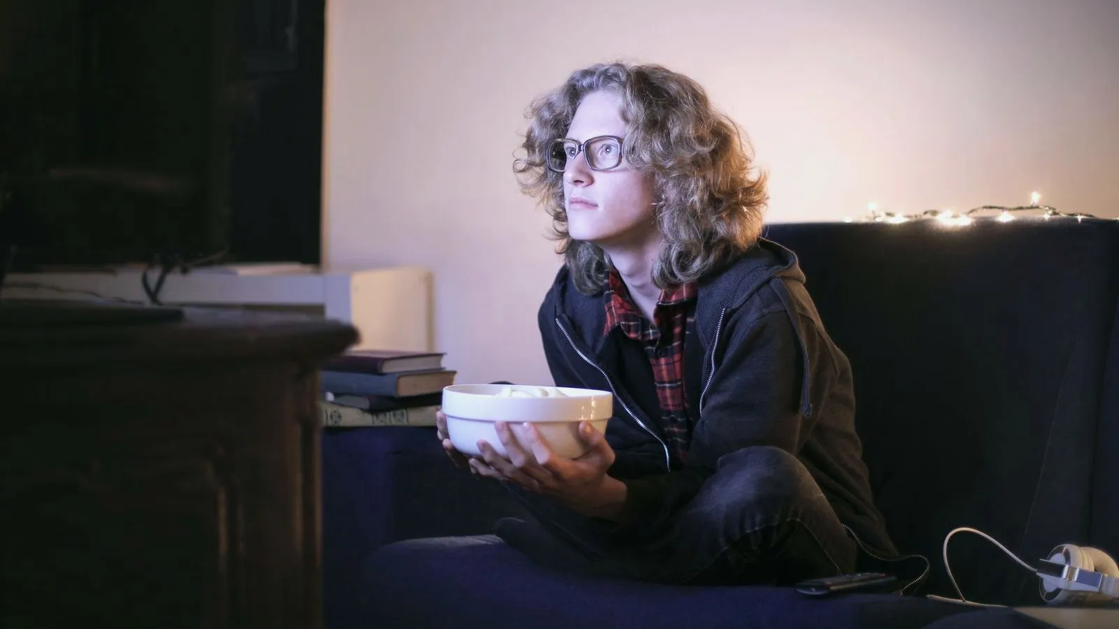 A person sitting cross-legged on a sofa at night holding a bowl, lit by a television