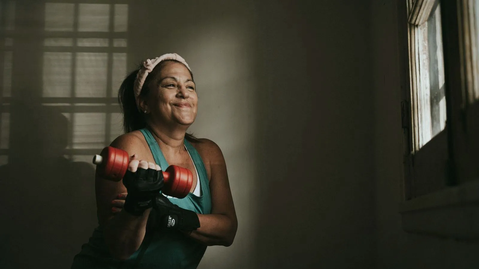 An older woman in a workout top and lifting glove smiles while raising a red dumbbell