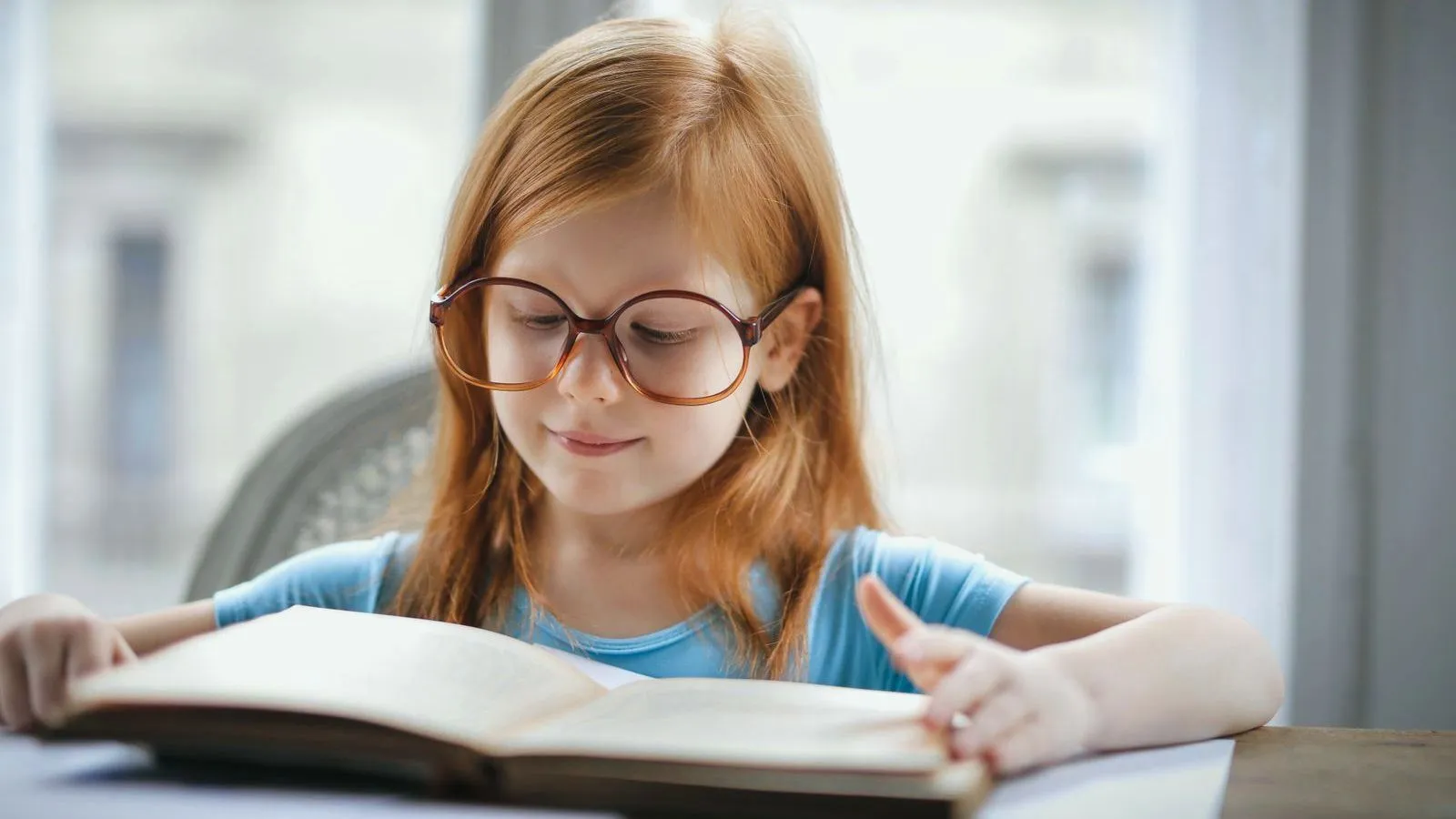 A young girl in round glasses reading an open book at a table