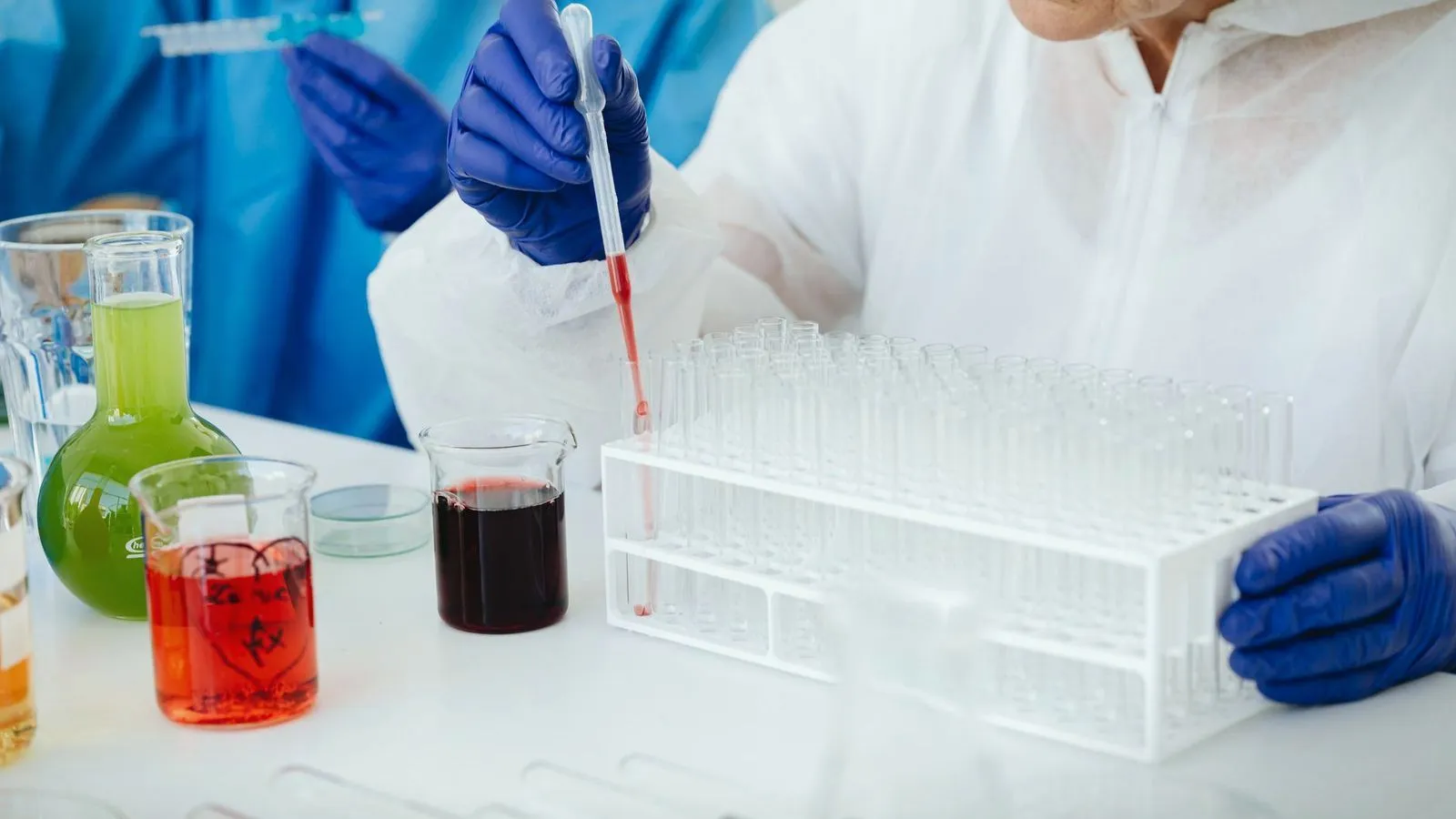 A gloved scientist pipetting a red sample into a rack of test tubes