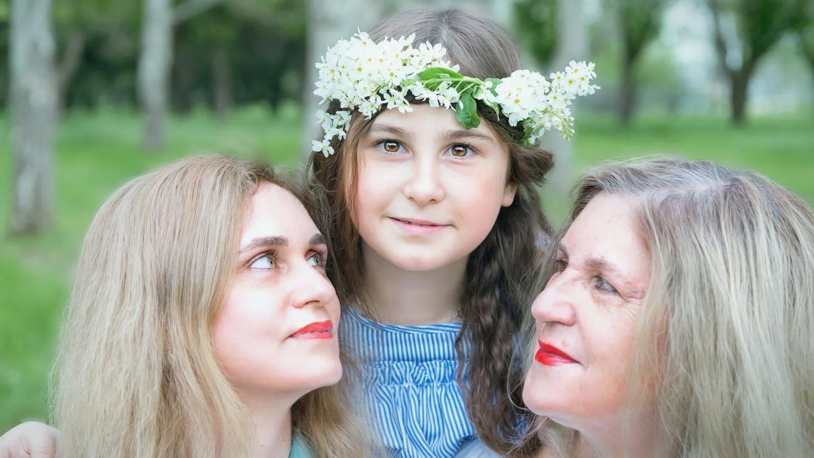 A girl wearing a crown of white flowers between a younger woman and an older woman