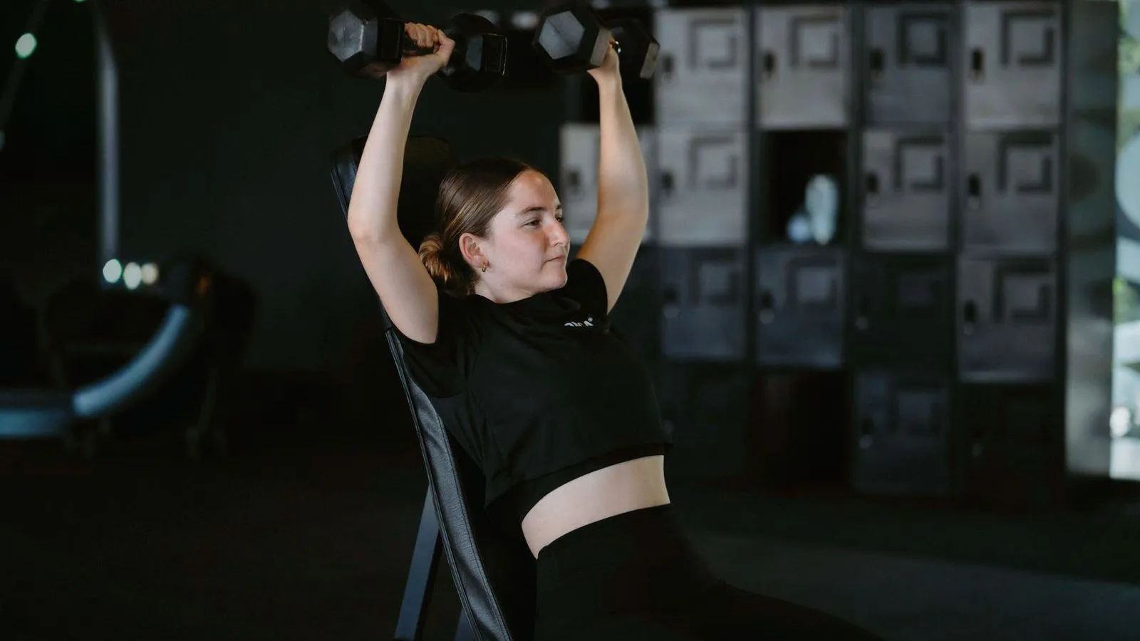 A woman pressing a pair of dumbbells overhead on a gym bench