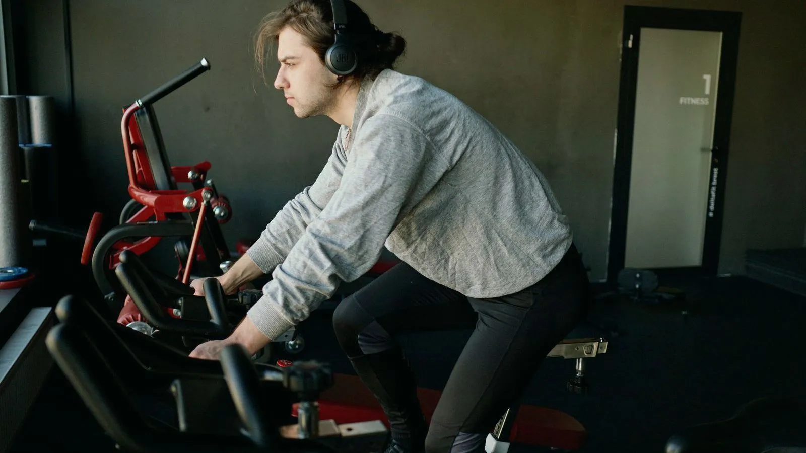 A young man in a grey sweater riding a stationary bike in a dimly lit gym