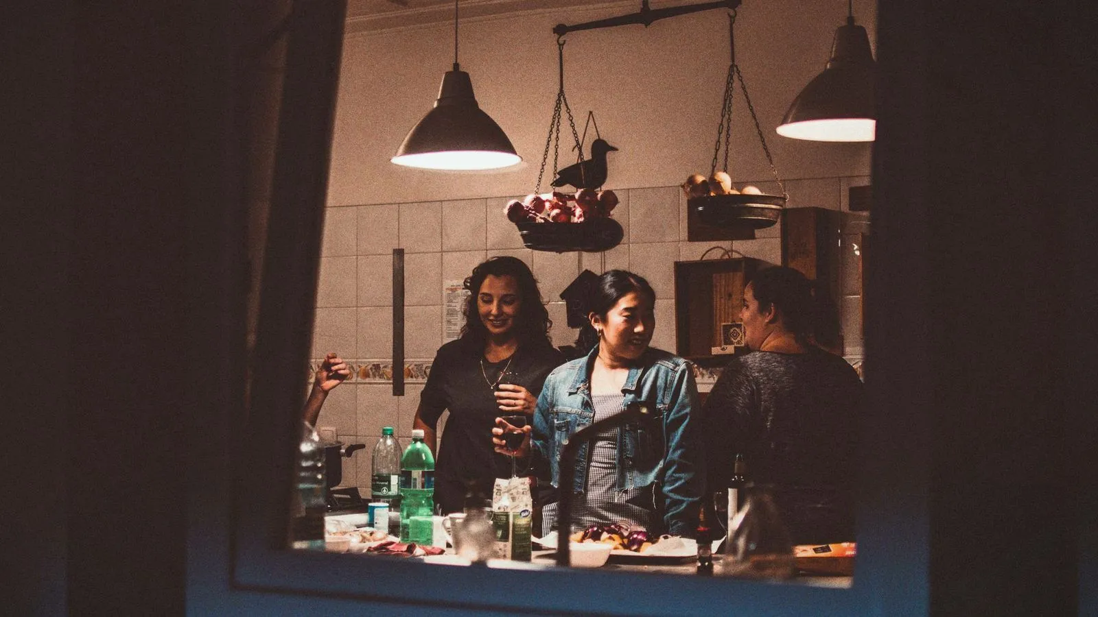 Three friends talking in a brightly lit kitchen late at night, seen through a doorway