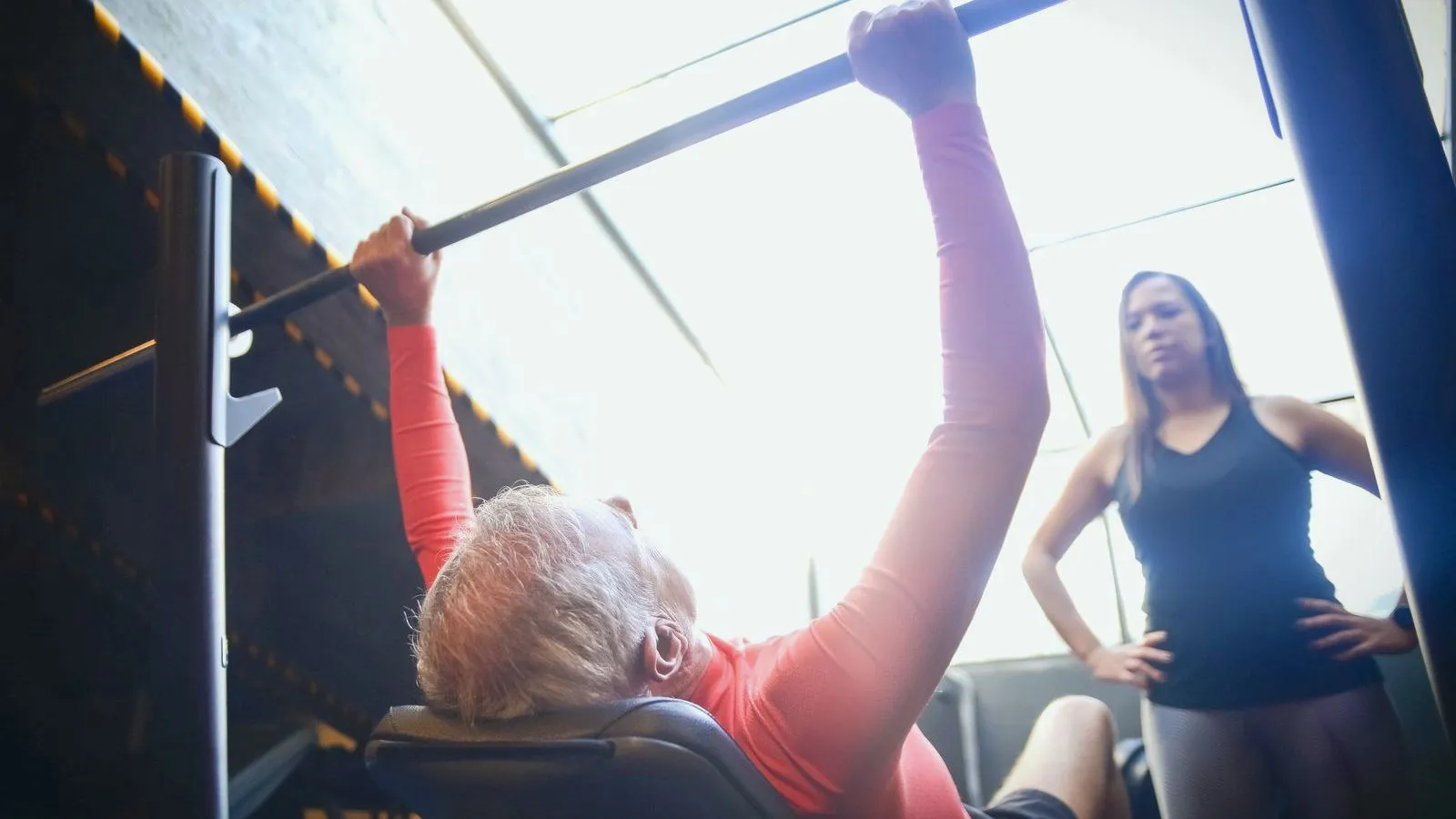 An older person bench pressing a barbell while a spotter stands over them