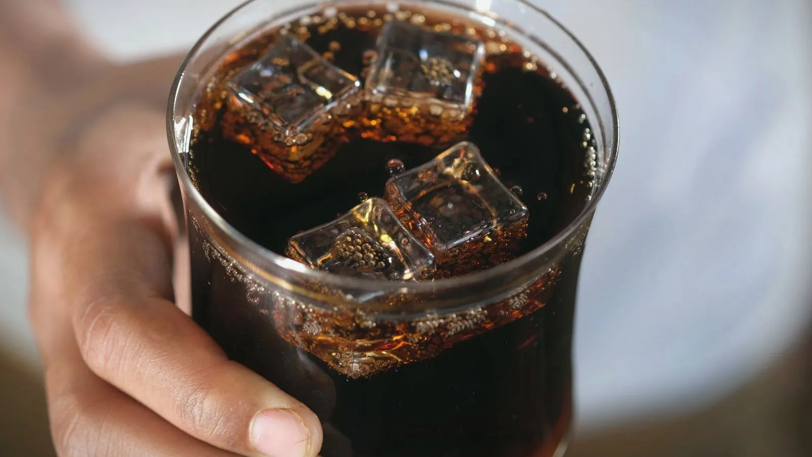 A hand holding a glass of dark cola filled with ice cubes