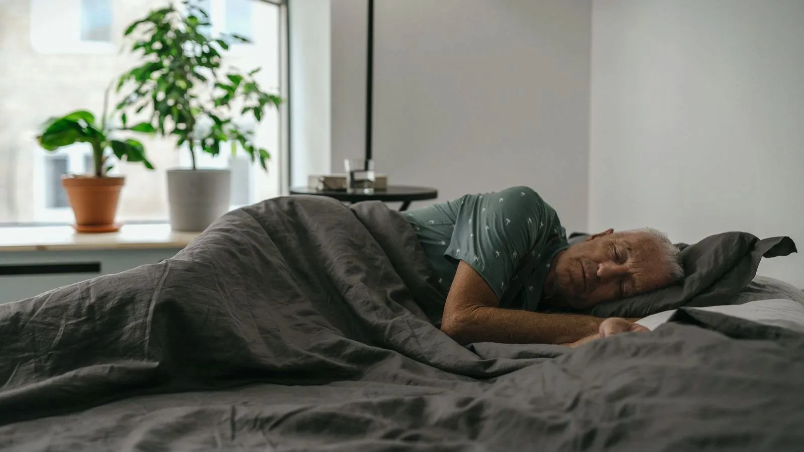 An older man asleep on his side in a bed beside a window with house plants