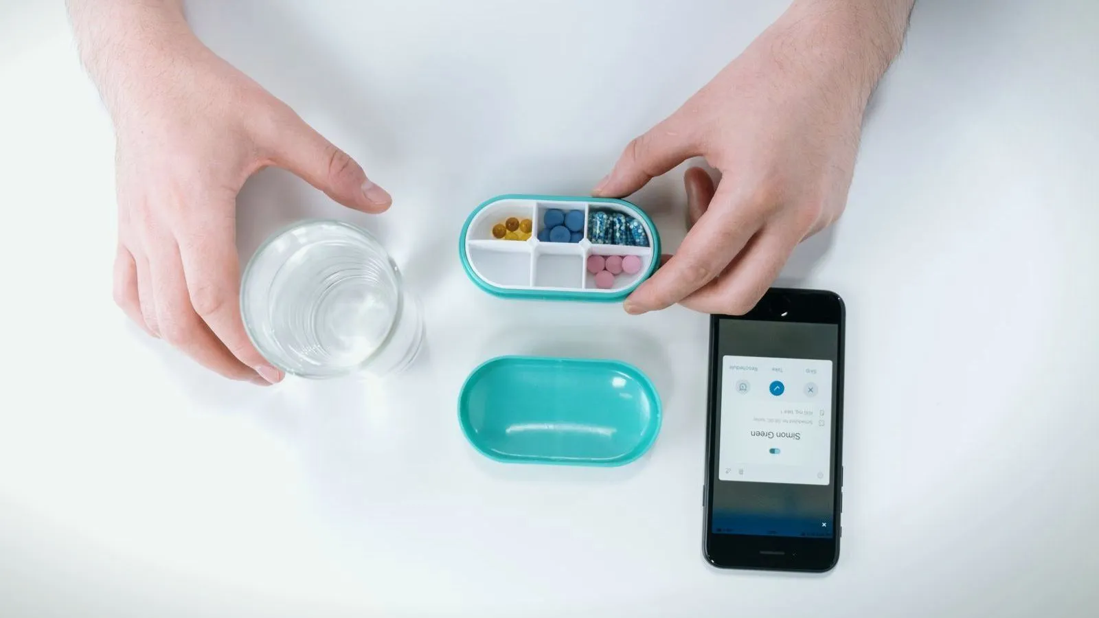 Hands filling a pill organizer beside a glass of water and a phone showing an app
