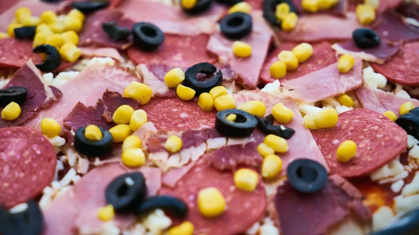 Close-up of an uncooked pizza topped with sliced ham, salami, black olive rings and sweet corn over grated cheese