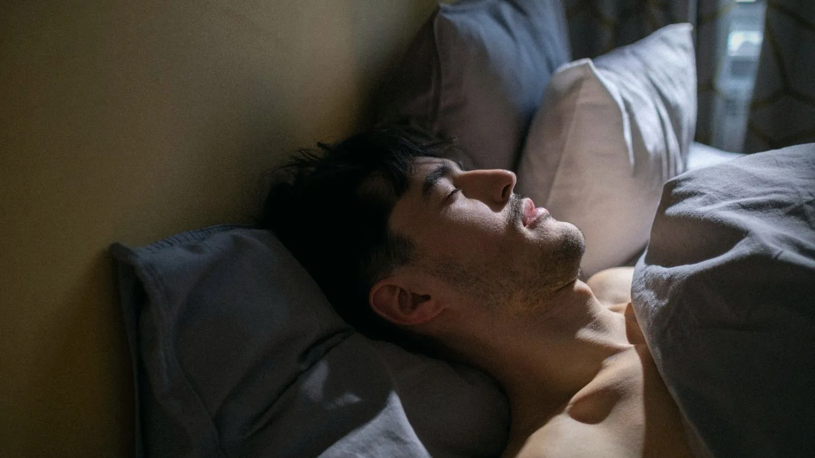 A man asleep on his back in a dim bedroom with his mouth open