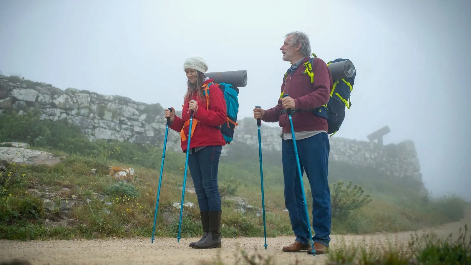 A woman and a gray-haired man with trekking poles and loaded backpacks standing on a misty hillside trail